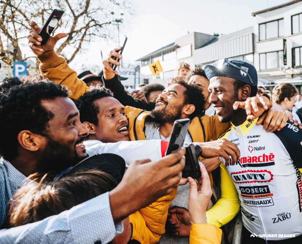 Group of men trying to take a selfie with an African cyclist in a yellow-and-white uniform