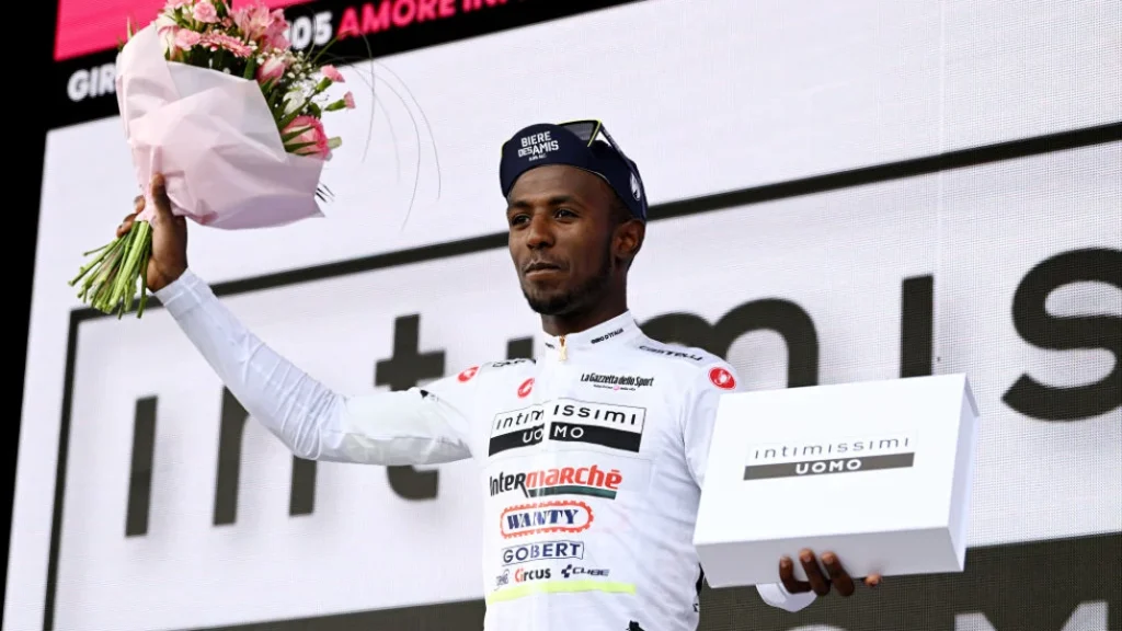 African man in a white cyclist uniform holding up a bouquet of pink and white flowers